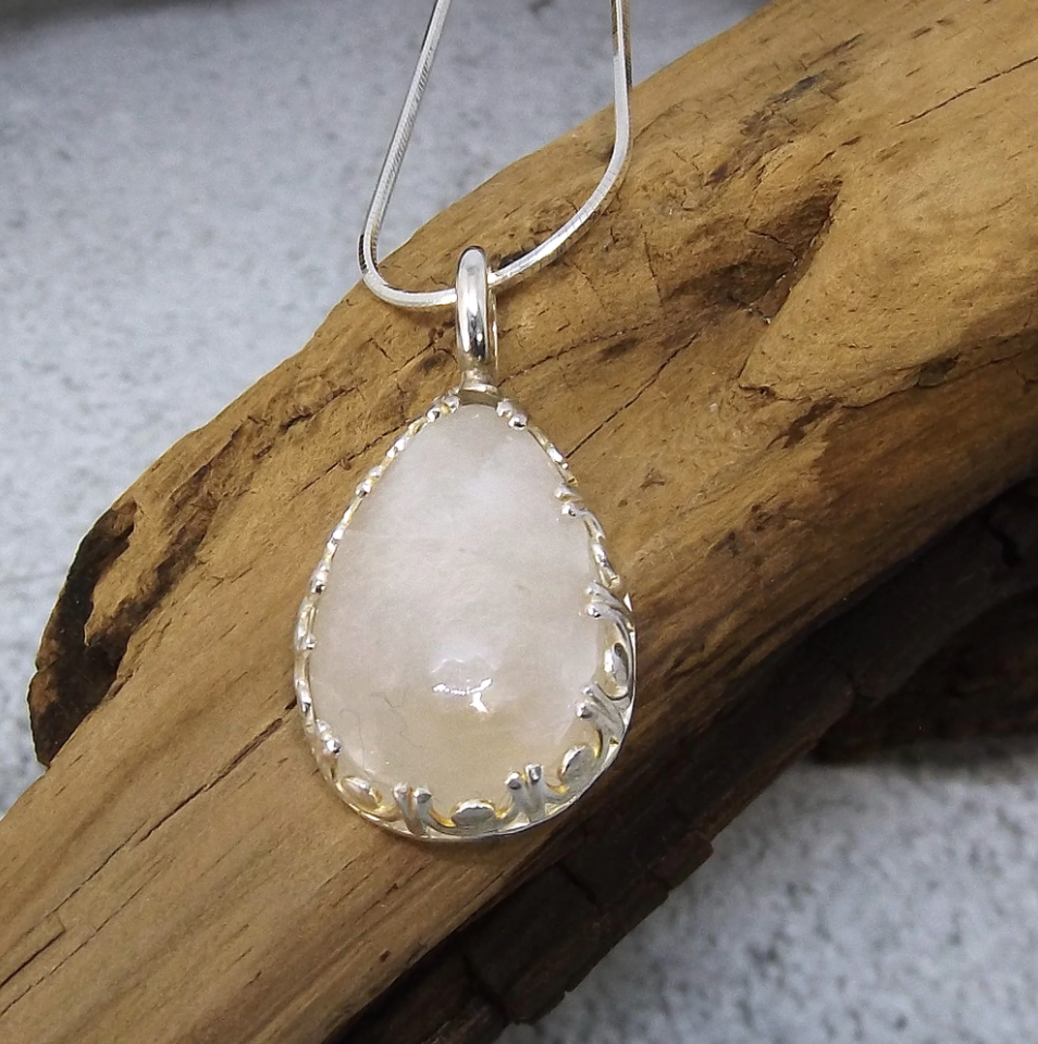 Screenshot of a photo of a necklace with a large whitish teardrop-shaped stone displayed on a piece of driftwood. 